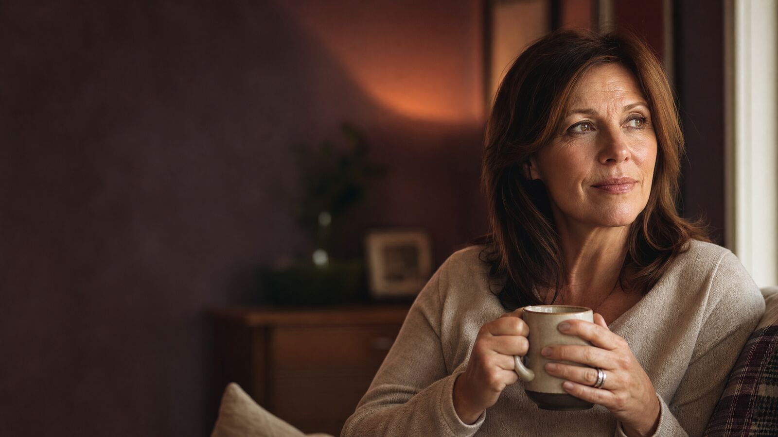 Woman in her late forties holding a ceramic mug, looking out a sunlit window in a quiet morning room